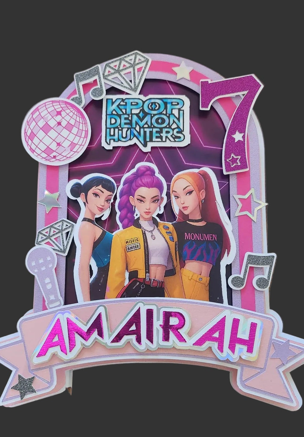 A decorative K-pop themed party decoration featuring characters from 'K-pop Demon Hunters' and the name on a pink and white banner